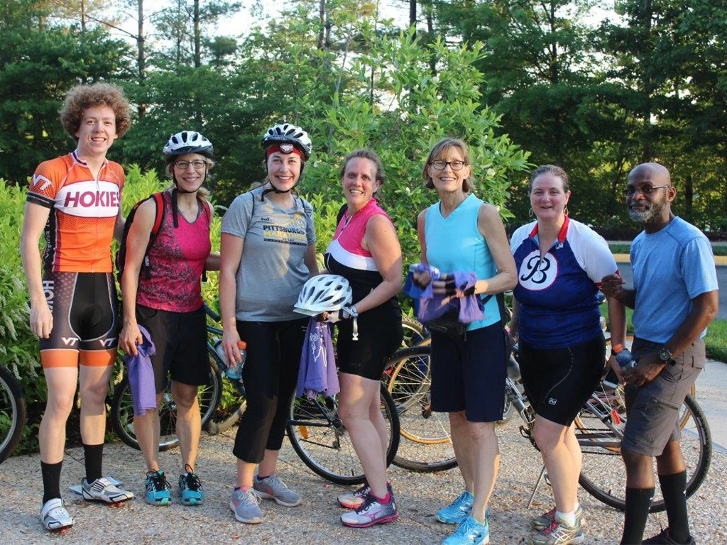 A group of seven adults, six women and one man, stand outdoors with their bicycles, smiling at the camera. They are wearing cycling gear and helmets, with trees and greenery in the background.