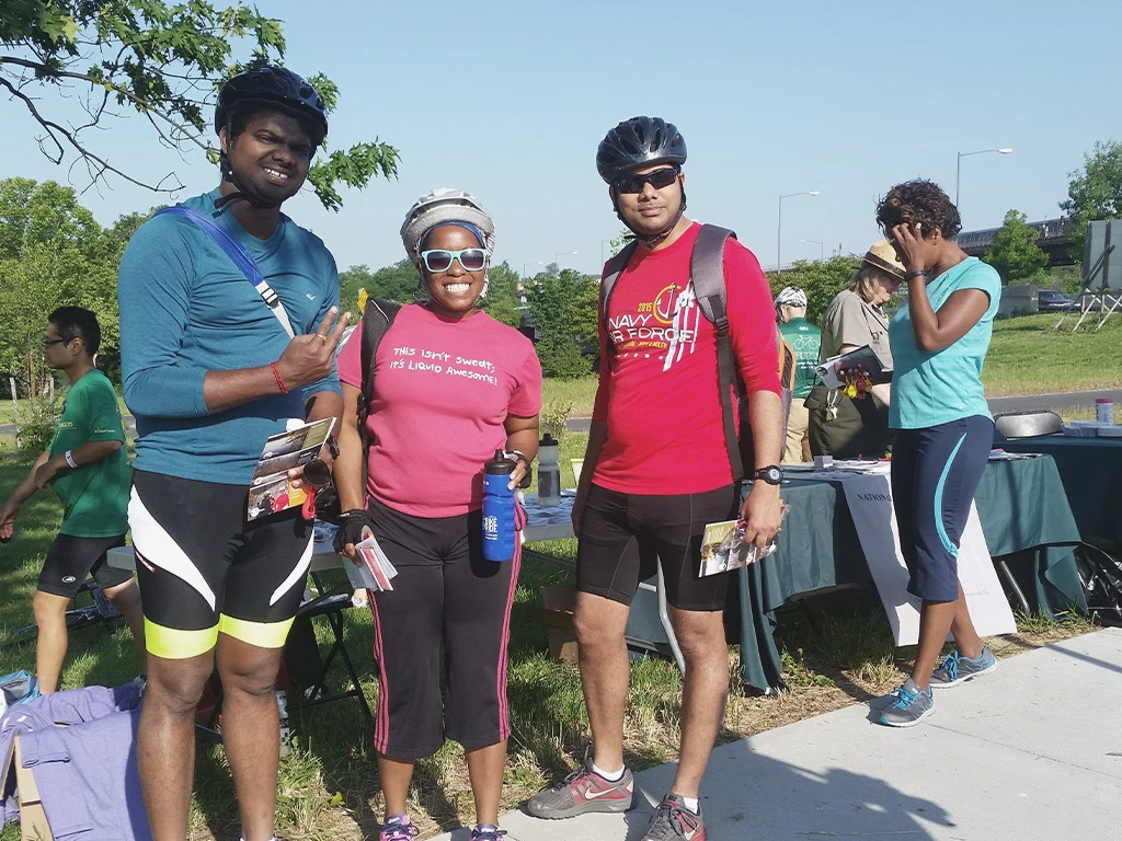 Three cyclists in helmets and athletic wear pose and smile outdoors at a park event, holding papers. Two other people stand at a nearby table under trees, with a blue sky and greenery in the background.