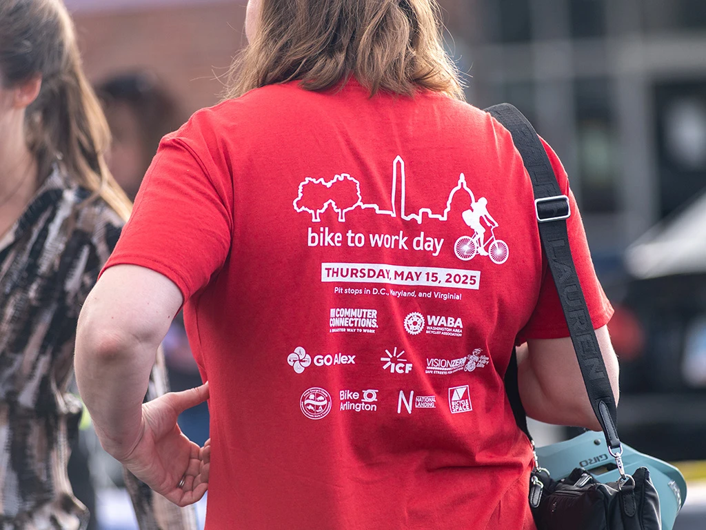Person wearing a red "Bike to Work Day" event T-shirt with white event details and logos on the back, standing outdoors among other people. The shirt displays the date May 15, 2025.