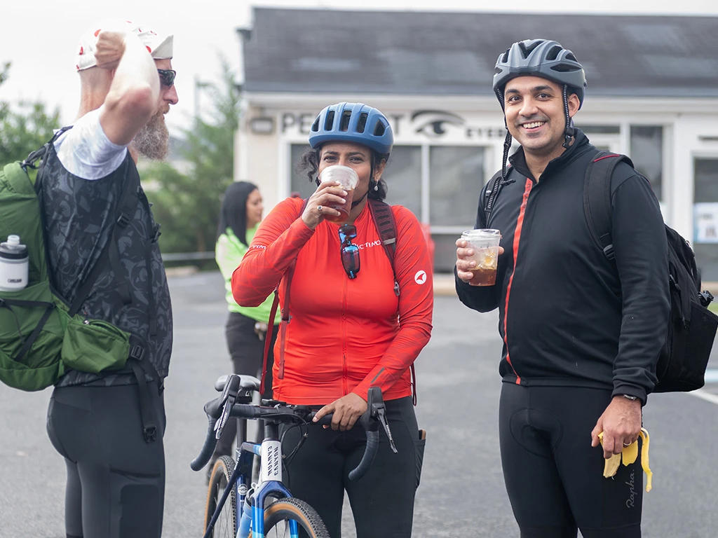 Three cyclists in helmets and cycling gear stand together outdoors. One woman holds a bike and drinks from a cup, while the two men stand nearby, one holding a banana. All appear relaxed and smiling.