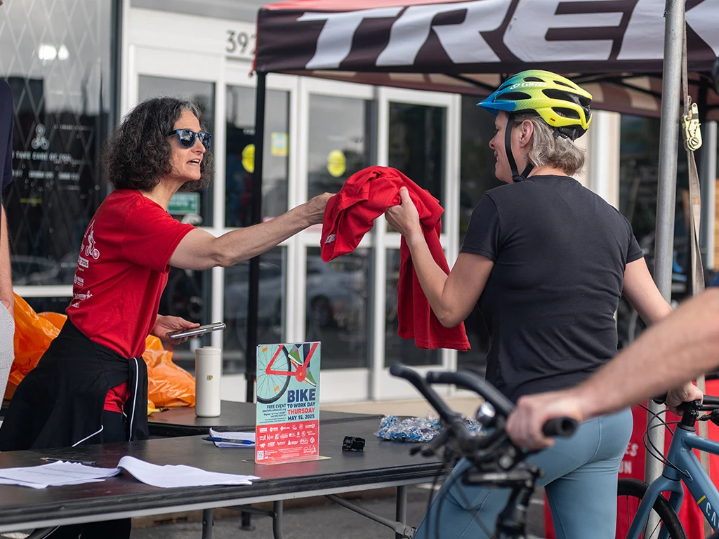 A woman in a red shirt hands a red t-shirt to a cyclist wearing a helmet at an outdoor event booth. A bike, a flyer, and other event materials are on the table between them.
