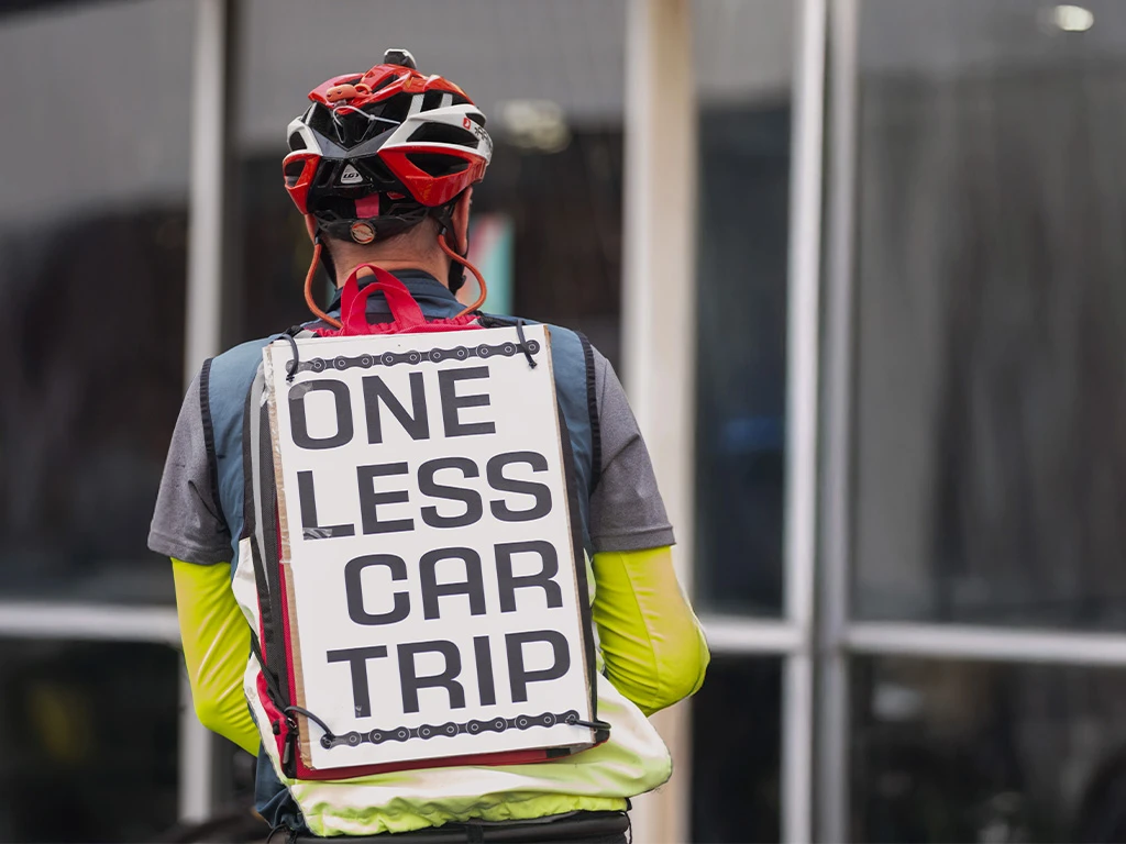A cyclist wearing a helmet and high-visibility clothing has a large sign on their back that reads, "ONE LESS CAR TRIP," as they stand near a building with glass windows.