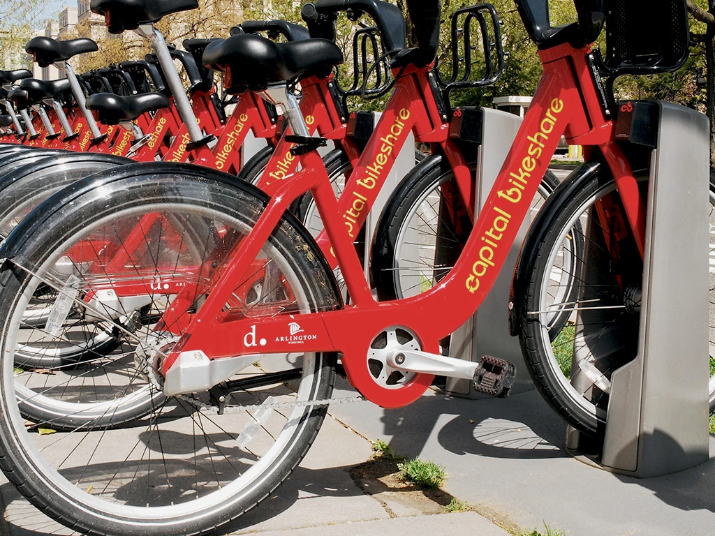 A row of red Capital Bikeshare bicycles is docked at a bike-sharing station on a sunny day, with the bikes lined up closely together, ready for public use.