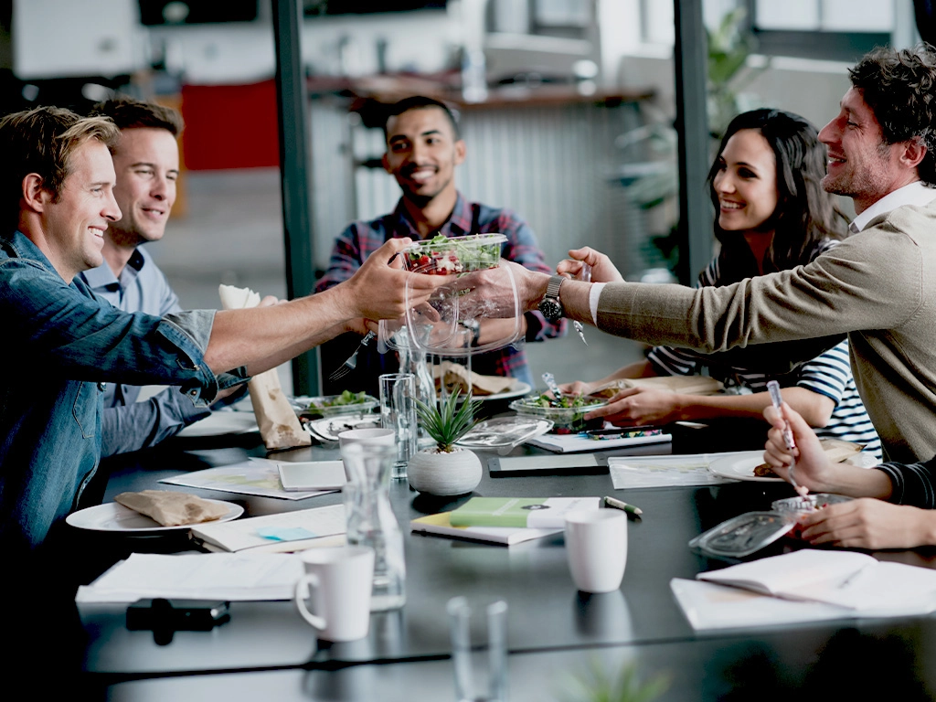 A group of six people sit around a table, smiling and sharing a meal together. They appear to be in a modern office setting, with notebooks, plates, and drinks on the table.