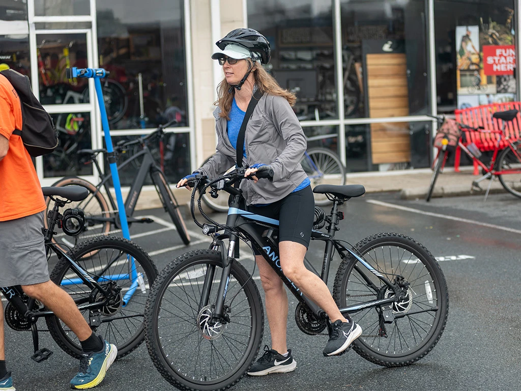 A woman wearing a helmet, sunglasses, gray jacket, and blue shirt stands astride a black mountain bike in a parking lot outside a bike shop. Several bikes and another person are visible nearby.