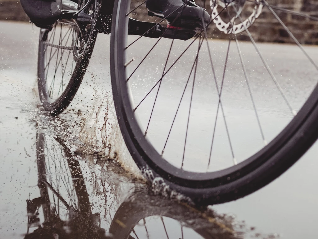 Close-up of a bicycle wheel splashing through a puddle on a wet road, with water droplets spraying and a clear reflection of the wheel visible in the water.