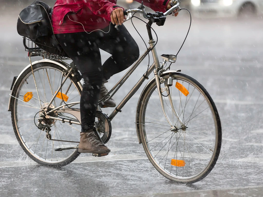 A person wearing a red jacket and brown boots rides a bicycle through heavy rain on a wet city street, with water splashing around the wheels.