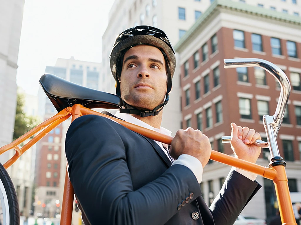 A man in a suit and helmet carries an orange bicycle over his shoulder while standing on a city street, with tall buildings in the background.