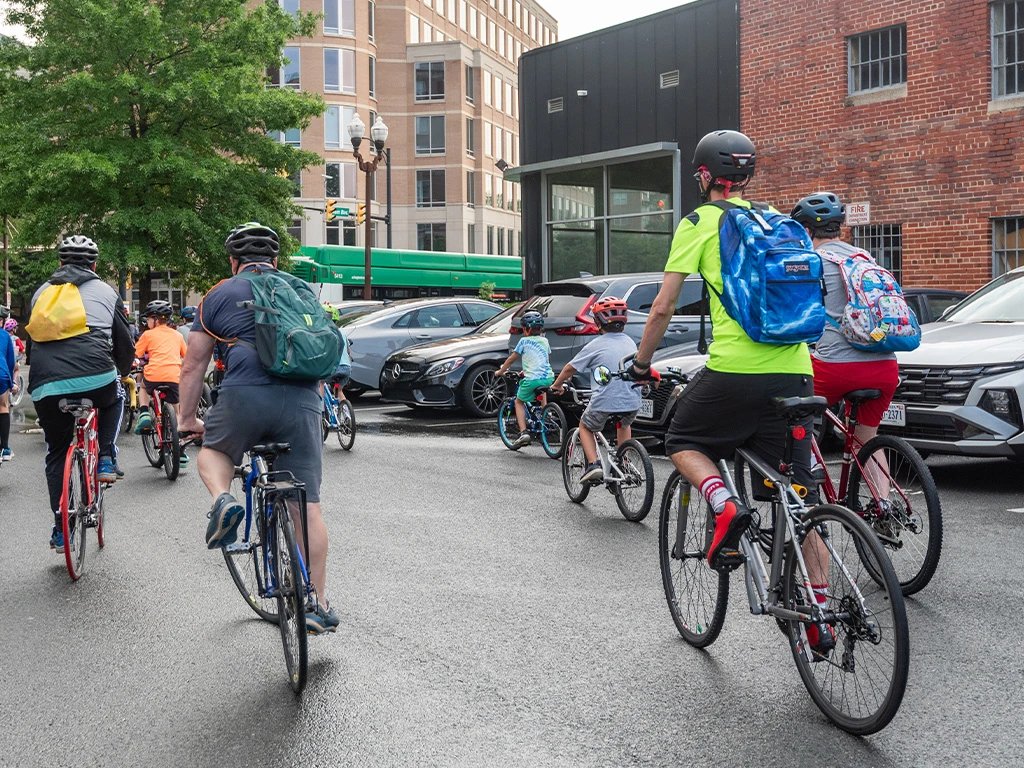 A group of cyclists, including adults and children wearing helmets and backpacks, rides together on a city street near parked cars and brick buildings. Trees and an apartment building are visible in the background.