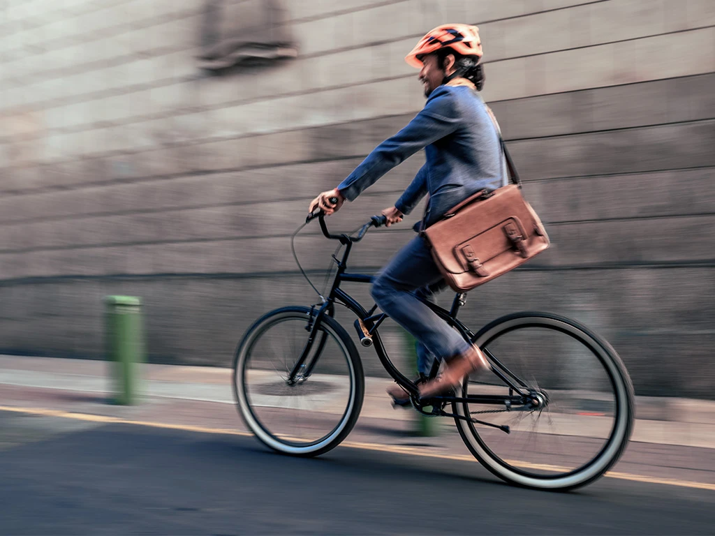 A person wearing a helmet and casual clothes rides a bicycle on a city street, carrying a brown shoulder bag. The background is blurred, indicating motion.