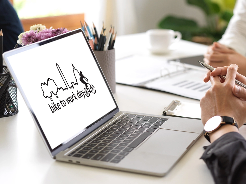 A person sits at a desk with a laptop displaying a "bike to work day" graphic, featuring a city skyline, trees, and a cyclist silhouette. Office supplies and papers are visible in the background.