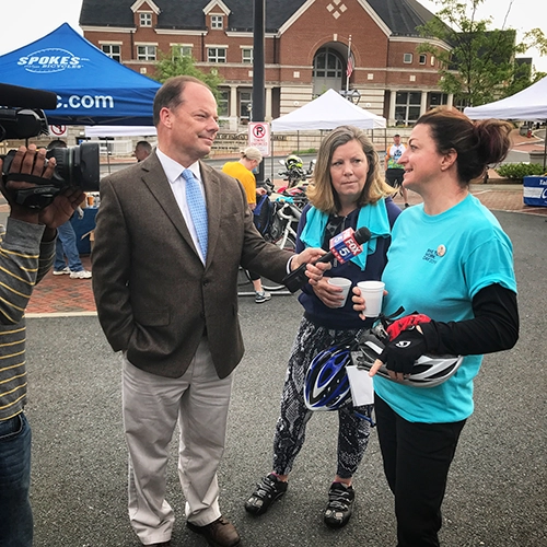 A news reporter interviews two women in teal shirts holding bike helmets at an outdoor event, while a cameraman films. Tents and a brick building are visible in the background.